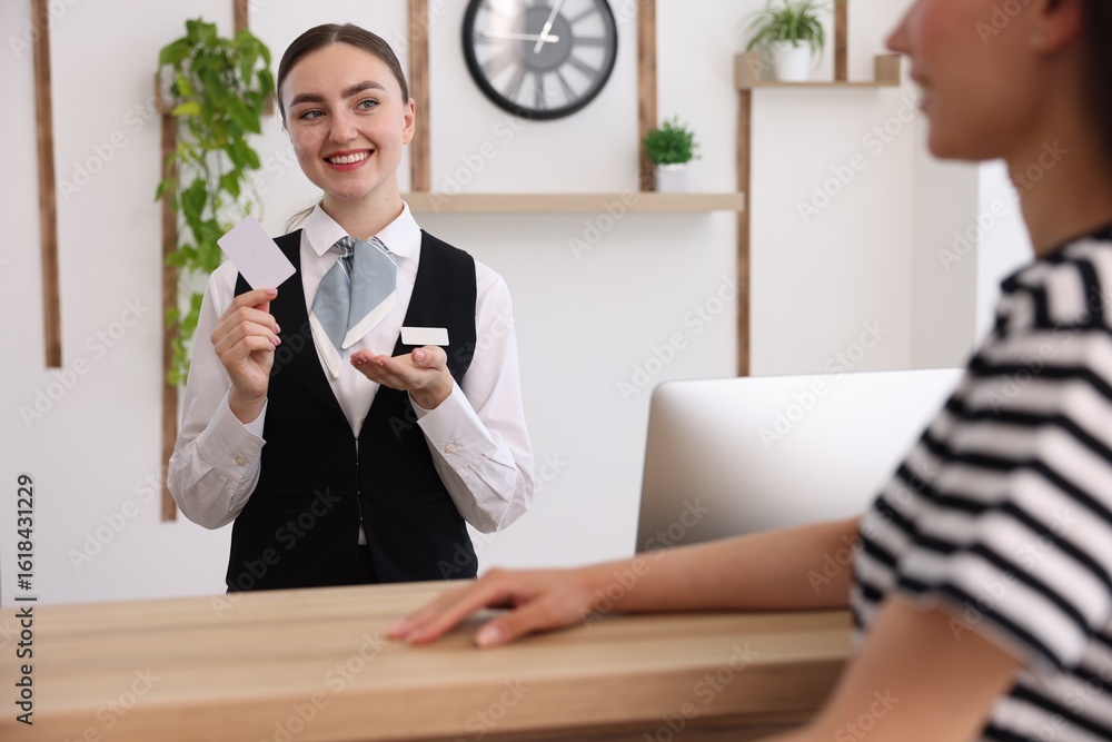 Poster smiling receptionist in uniform with hotel key card working with guest at reception desk indoors
