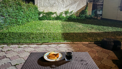 Night snack on outdoor table in backyard garden. A plate with baked pastries and a glass of dark soda on a wicker-style table in a backyard at night. The yard is lit with artificial light.