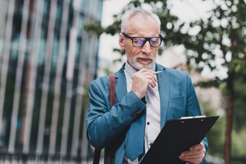 Mature businessman in formal suit reviewing documents outdoors in urban setting, emphasizing...