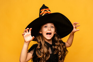Happy funny child girl in witch costume and black hat makes horror face and grimaces on yellow...