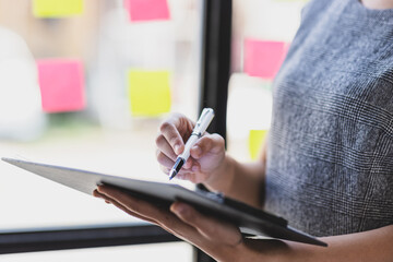 Creative Asian businesswoman thinking and planning startup business with paper stickers on glass, analyzing strategy and planning business in office.