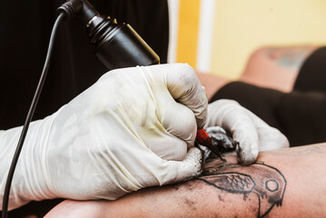 Gloved tattooist works on bird skull tattoo with red cartridge, outlining and adding shading to...