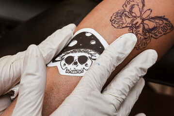 Tattoo artist wearing white gloves places a mushroom skull stencil next to an already transferred...
