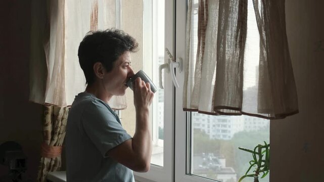 Middle-aged woman with short hair holding a coffee cup and standing by the window at home, enjoying a calm moment in the morning light.