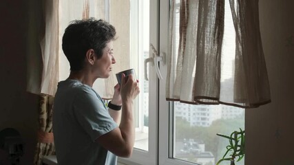 Side view of a middle-aged woman with short hair holding a cup of coffee and looking out the window, capturing a moment of relaxation and daily routine at home - Powered by Adobe
