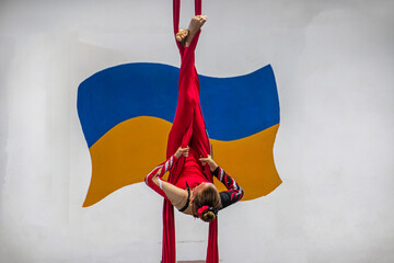 A beautiful aerial gymnast in a red costume hangs against the backdrop of the Ukrainian flag	
