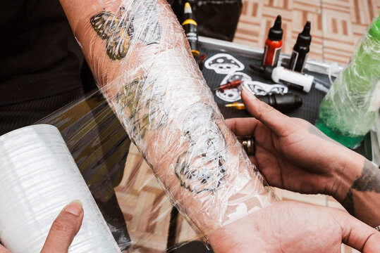 Tattoo artist covers newly finished forearm tattoos with transparent plastic wrap to protect skin and preserve ink during healing.