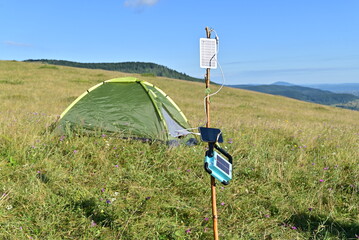 Smartphone charging via portable solar panels beside a green tent in a sunny mountain meadow.