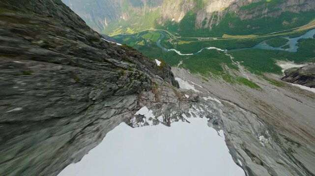 Aerial view of steep gray cliffs meeting green fields bisected by a winding river, with patches of white snow clinging to the rock face, Stranda Municipality, Norway.