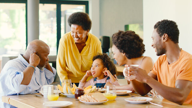 Family Shot With Grandparents Parents And Granddaughter Pulling Faces At Breakfast Around Table At Home