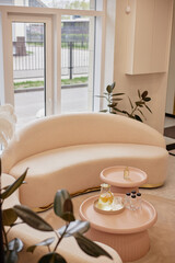 Curved pastel sofa placed beside large window with soft natural light filling modern waiting room featuring small round tables holding refreshment drinks and fragrance bottles
