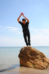 A guy in a black suit stretched out in a jump from a rock onto a sandy seashore against the...