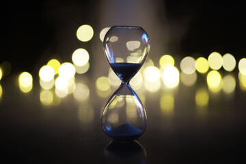 Glass Hourglass with Blue Sand and Bokeh Lights Background: Symbol of Time and Transience Concept