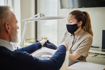 Caucasian middle aged man receiving manicure from young Caucasian woman wearing protective mask,...