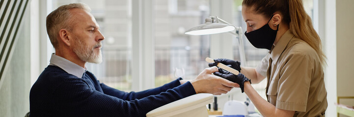 Website header shot of Caucasian middle aged man receiving manicure from young Caucasian woman wearing protective mask and gloves, man sitting at table while woman filing his nails in modern salon