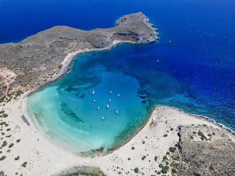 Aerial view of the serene turquoise waters meet the pale sands, cradling boats under the watchful gaze of rocky cliffs, Elafonisos, Greece.