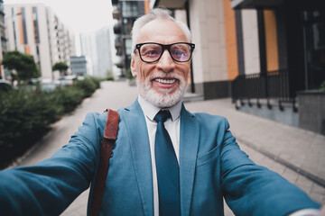 Successful senior businessman taking a cheerful selfie outdoors in an urban modern city street...