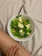 Realistic moment of eating a salad in bed — mozzarella, greens, and casual setting reflect...