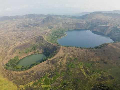 Aerial view of the contrasting deep blue and murky green lakes nestled in volcanic craters, surrounded by dry, golden grasslands and distant mountains, Nkomgsamba, Southwest Region, Cameroon.