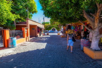 Shops on cobblestone pedestrian street with woman walking, selling various tourist souvenirs and Mexican crafts, backs to camera, sunny day in Loreto, Baja California Sur, Mexico