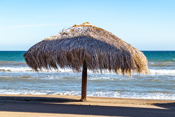 Palapa on beach in coastal landscape, waves breaking on shore and sea in background, brown sand, sunny spring day in Loreto, Baja California Sur, Mexico