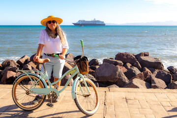 Female tourist standing next to bicycle on boardwalk, sea and cruise ship in background, wearing shorts, white blouse, hat and sunglasses, sunny day in Loreto, Baja California Sur, Mexico