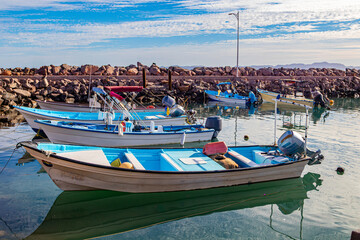 Fishing motor boats anchored on calm waters of Loreto Harbor Bay, boardwalk with breakwaters in background, cloudy day in Baja California Sur, Mexico