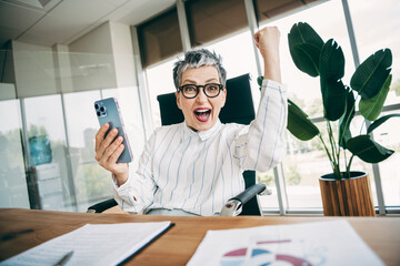 Excited businesswoman celebrating success in office holding phone at desk with charts and plants...