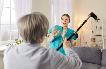 Angry elderly senior woman going to hit young female nurse with crutch. Enraged exasperated female...