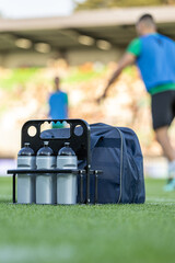 Water bottles and sports bag on a soccer field sideline during training. Concept of hydration, refreshment, and team sports with blurred players on the green grass © dechevm