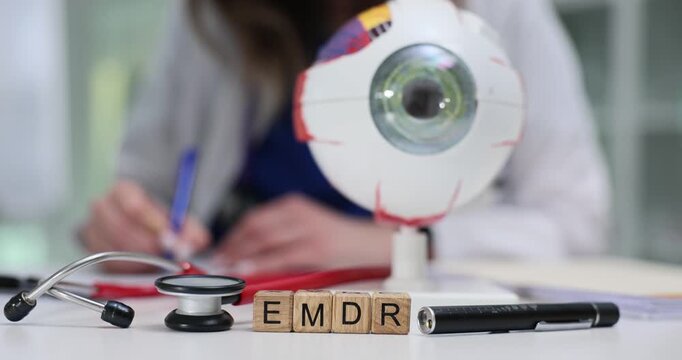 Wooden blocks with EMDR and model of eyeball on desk near doctor writing records. Focus on eye movement desensitization and reprocessing therapy