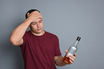 Man with bottle of whiskey suffering from hangover on grey background