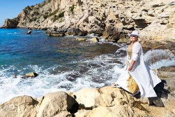 A woman in a long dress with a flowing white cape and a white hat stands among rocks on the shore...