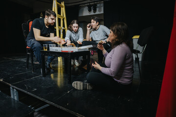 A group of actors sharing a meal during a rehearsal in a theatre setting. The warm atmosphere conveys teamwork and camaraderie.