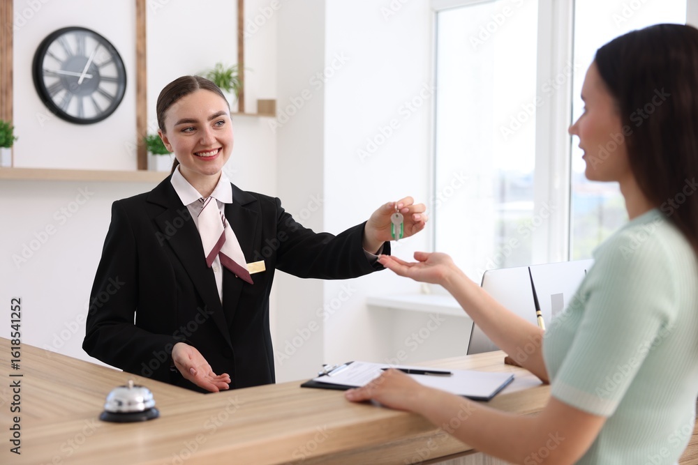 Poster smiling receptionist in uniform giving guest room key to client at reception desk in hotel