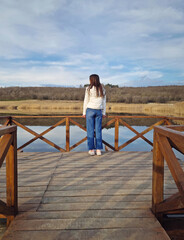 Rear view carefree young woman stands on a wooden pier over calm body of water, surrounded by dry reed vegetation. Rustic vibe on the footbridge with a lone girl enjoying the beauty of nature