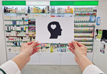 Pharmacist hands holding a paper with a head silhouette featuring a heart inside, symbolizing mental health and wellness, over pharmacy shelves backdrop. Physical self-care and emotional well-being