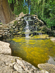 Graceful spring with cascades over moss-covered stones into a crystal-clear pond with thrown coins. Golden sunlight dances on the fountain rippling water. The serene scene radiates natural beauty