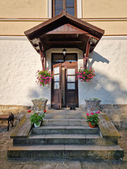 Rustic old building facade with porch and a charming wooden door, framed by two side windows and a small overhang. Stone steps lead up to the entrance, adorned with flower pots in bloom