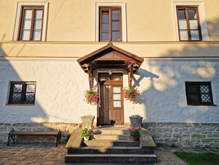 Rustic old building facade with porch and a charming wooden door, framed by two side windows and a small overhang. Stone steps lead up to the entrance, adorned with flower pots in bloom