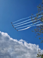Air show with five airplanes flying in formation with white smoke trails curving upward through a bright blue sky scattered with fluffy clouds