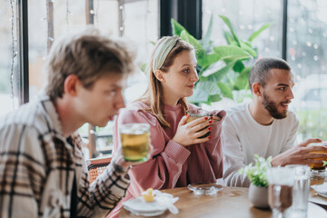 Three friends sit around a table enjoying warm drinks, creating a cozy and sociable environment with a focus on camaraderie and relaxation in a comfortable cafe atmosphere surrounded by greenery.