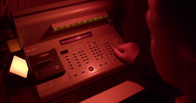 An artist at work in the atmospheric red glow of a photo darkroom. A hand is seen programming an exposure on a vintage enlarger console.