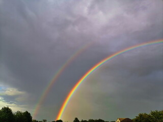 A bright double rainbow arches across a dramatic dark cloudy sky above a green forest, creating a...