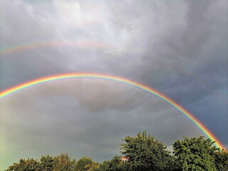 A vivid double rainbow glows brightly across the cloudy evening sky above lush green trees,...