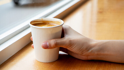 Person holding a disposable coffee cup near a window on a wooden table