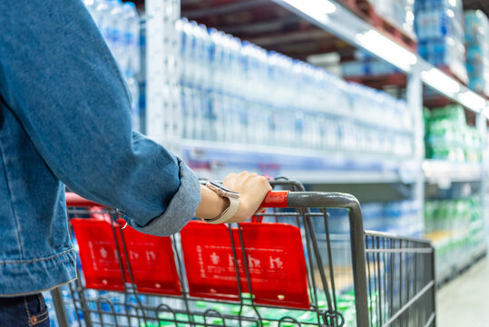 Image of asian woman shopper pushing a red shopping cart in a warehouse store aisle, surrounded by shelves full of bottled water and other items ready for customers to add to their carts.