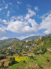 A scenic view of a mountain village in Nepal, featuring traditional houses nestled on a hillside under a blue sky with fluffy clouds. Ideal for travel, tourism, or cultural heritage content.