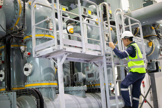 Technician performs maintenance on gas insulated switchgear inside substation wearing safety gear and helmet while climbing metal ladder to access equipment