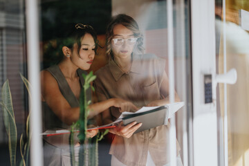 Two women are viewed through a glass window, focusing on a document in a collaborative and...